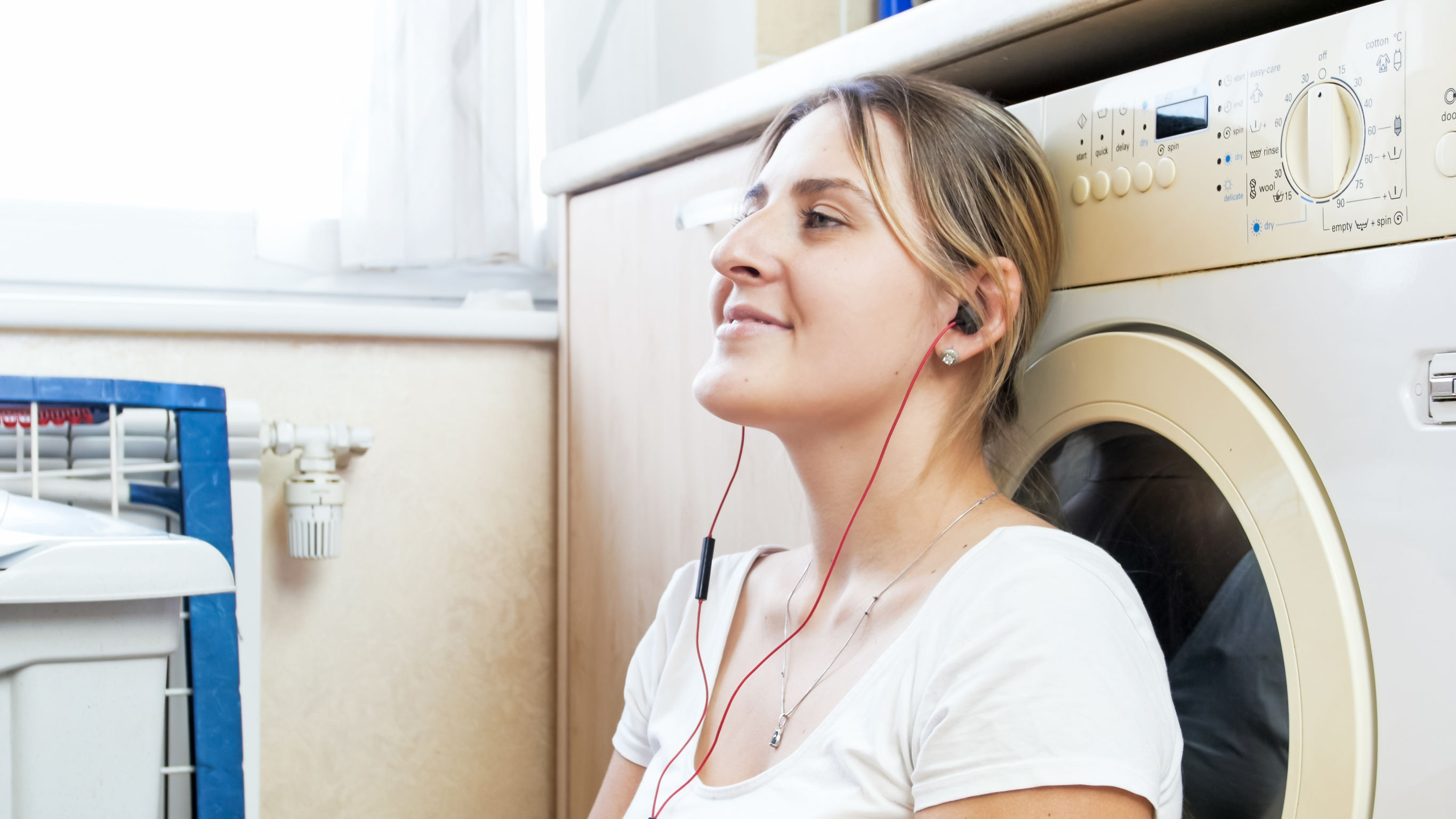 Portrait of beautiful housewife with earphones leaning on washing machine at laundry